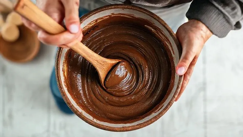 A photorealistic, professional photography shot from above, showing hands gently mixing a rich, dark brown millet brownie batter in a large ceramic bowl with a wooden spoon. The batter has a smooth, creamy texture. Cinematic lighting, sharp focus on the mixing action, depth of field blurring the background of a clean, minimalist kitchen. 8K, shot on a high-end DSLR, conveying a sense of homemade goodness and culinary precision.