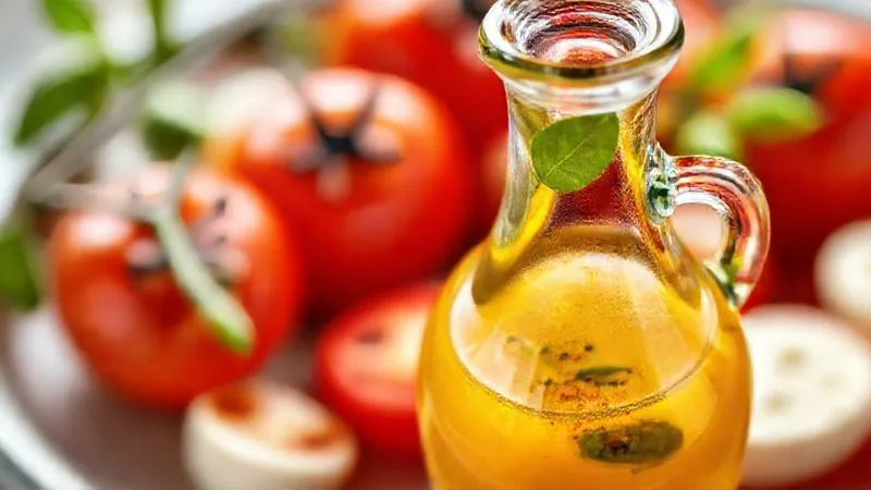 A photorealistic, professional photography, 8K, cinematic lighting, sharp focus, depth of field image of a light, vibrant Caprese dressing. A small, elegant glass cruet holds a clear, golden liquid with a hint of red, suggesting a blend of balsamic glaze and lemon juice, with fresh basil leaves floating within. The background shows blurred, fresh tomatoes and mozzarella, emphasizing the healthy and flavorful dressing. Shot on a high-end DSLR, highlighting the freshness and low-calorie appeal.