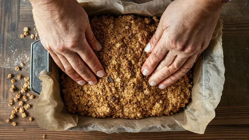 A top-down, photorealistic, professional photography shot of hands firmly pressing a granola bar mixture into a parchment-lined baking dish. The mixture looks dense and slightly sticky. Cinematic lighting highlights the texture of the ingredients, sharp focus, depth of field, 8K hyper-detailed, shot on a high-end DSLR.