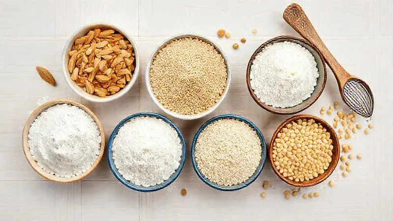 A vibrant flat lay of various gluten-free flours: almond flour, coconut flour, full-grain rice flour, and chickpea flour, arranged in small bowls on a light wooden background, with measuring spoons and a whisk. Photorealistic, professional photography, 8K, cinematic lighting, sharp focus, depth of field, shot on a high-end DSLR.