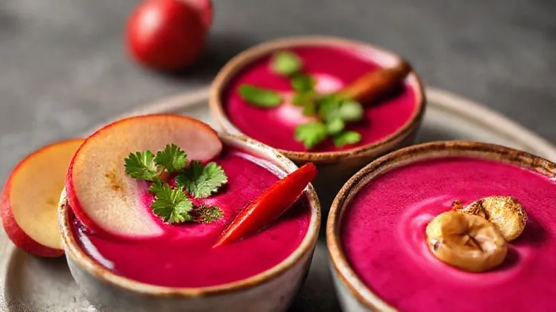 Photorealistic image of three small, distinct bowls of beet cream soup variations: one garnished with apple slices and ginger, another with fresh cilantro and a red chili, and a third with roasted garlic cloves, all arranged artfully, cinematic lighting, 8K hyper-detailed.