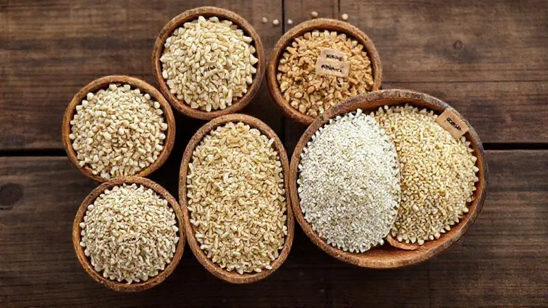Photorealistic, professional photography, 8K, cinematic lighting, sharp focus, depth of field, shot on a high-end DSLR. A close-up, top-down view of various types of uncooked dry grains arranged neatly in separate small wooden bowls: buckwheat, pearl barley, millet, quinoa, and bulgur. Each bowl is labeled with a small, elegant handwritten tag in Polish. The background is a rustic wooden table, softly blurred.