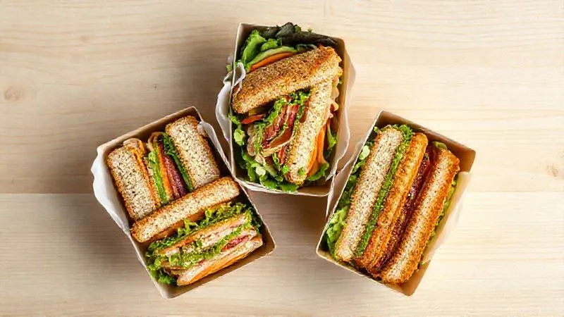 A photorealistic, top-down shot of three different, beautifully arranged full-grain sandwiches, each distinct and colorful, presented in bento-style lunchboxes on a light wooden table. Professional photography, 8K, cinematic lighting, sharp focus, depth of field.