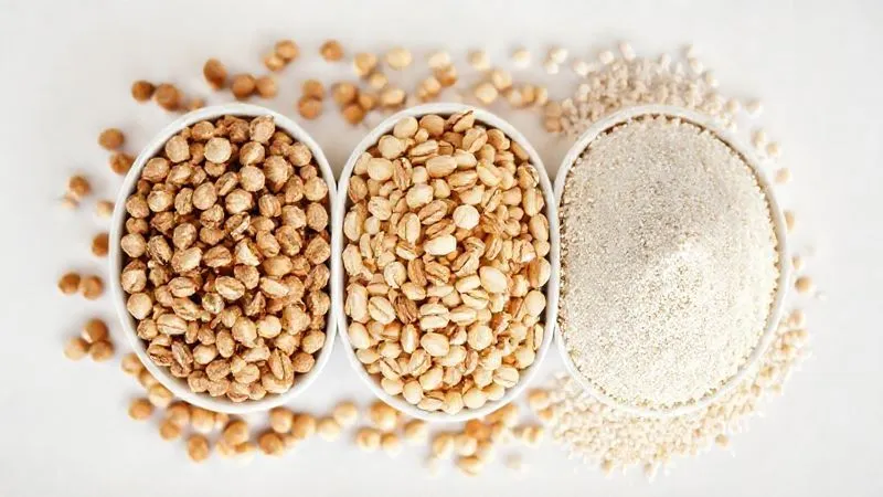 A professional photography image showcasing three distinct forms of buckwheat side-by-side: whole roasted buckwheat groats, whole unroasted (white) buckwheat groats, and buckwheat flour. Each type is in a small, elegant bowl, with a clean, minimalist background, cinematic lighting, sharp focus on the textures, 8K hyper-detailed, shot on a high-end DSLR.
