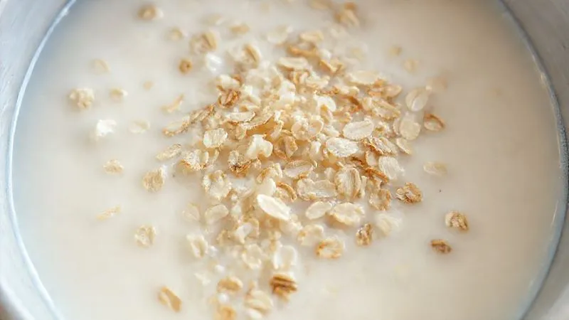 A close-up, photorealistic image of dry oat flakes soaking in a bowl of warm milk, with some flakes already plump and others still absorbing liquid, cinematic lighting, sharp focus on the texture change, 8K hyper-detailed.