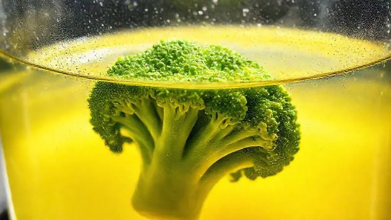 Photorealistic image of a transparent glass pot with boiling water, and a few pale, overcooked broccoli florets sinking to the bottom, surrounded by yellowish water, emphasizing nutrient loss. Cinematic lighting, 8K, sharp focus, depth of field.