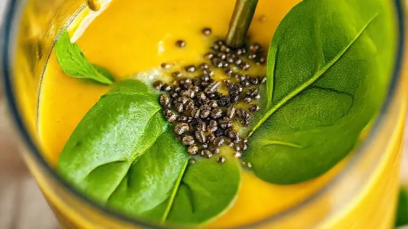 A photorealistic close-up of a high-speed blender in action, incorporating vibrant green spinach leaves and tiny chia seeds into a creamy yellow mango smoothie. The texture should look rich and inviting, symbolizing hidden nutrients and effective blending. Cinematic lighting, sharp focus on the blending ingredients, 8K hyper-detailed.