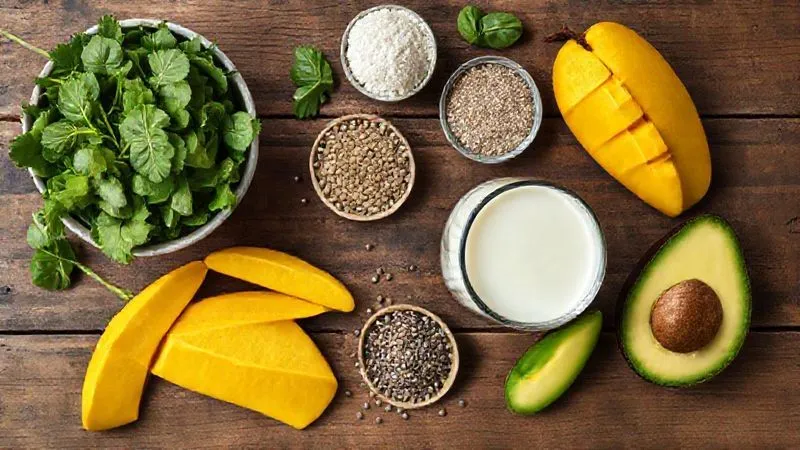 A photorealistic, top-down shot of all the fresh ingredients for a healthy, balanced mango smoothie beautifully arranged on a rustic wooden table. The ingredients include ripe mango slices, a bowl of fresh spinach, a small glass of unsweetened almond milk, a scoop of vanilla protein powder, a small pile of chia seeds, and a quarter of an avocado. The composition should be clean, inviting, and professional. 8K hyper-detailed, cinematic lighting, sharp focus on the ingredients.
