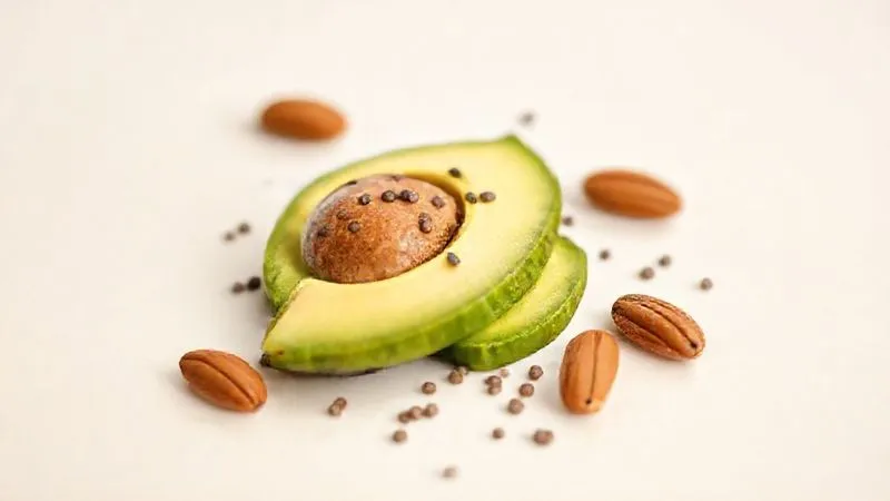 A close-up, photorealistic image of a small, artfully arranged portion of healthy fats: sliced avocado, a sprinkle of chia seeds, and a few whole almonds, on a light, minimalist background. Cinematic lighting, sharp focus, depth of field, 8K, shot on a high-end DSLR.