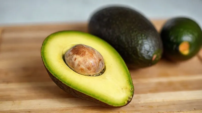 Photorealistic, professional photography of a perfectly ripe Hass avocado, cut in half, showcasing its creamy, light green flesh and a single pit. The avocado is placed on a clean wooden cutting board, with soft, natural light emphasizing its smooth texture. Depth of field, sharp focus on the avocado, 8K hyper-detailed, shot on a high-end DSLR.