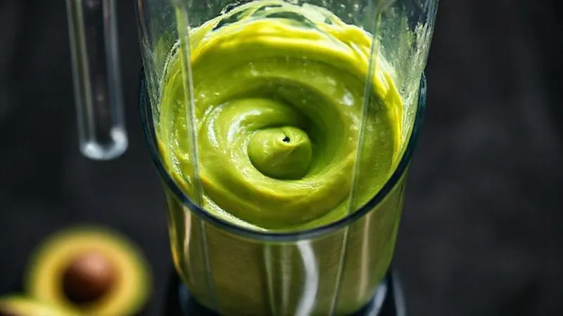 Photorealistic, professional photography of a high-speed blender actively blending a vibrant green avocado mixture, creating a smooth, swirling vortex. The blender jar is clean, and the focus is on the creamy texture being formed. Cinematic lighting, sharp focus, depth of field, 8K hyper-detailed, shot on a high-end DSLR.