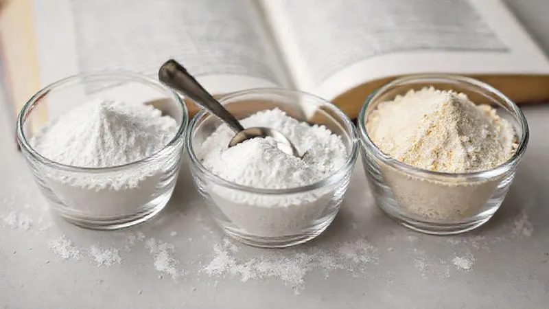 Photorealistic close-up of three small glass bowls, each containing a different white powdered stabilizer: agar-agar, xanthan gum, and cornstarch. Each powder is perfectly white and fine, with a small silver measuring spoon resting in each bowl. Cinematic lighting, sharp focus, depth of field blurring a cookbook in the background, 8K hyper-detailed, professional photography.