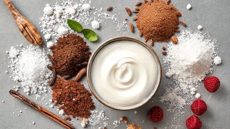 A vibrant photorealistic flat lay of various natural sweeteners and flavorings arranged around a small bowl of light coconut cream. Ingredients include erythritol crystals, a dropper of liquid stevia, cocoa powder, vanilla bean pods, and freeze-dried raspberry powder. Soft, natural light, sharp focus on all elements, professional food photography, 8K hyper-detailed.