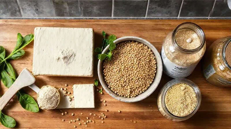 A vibrant, photorealistic overhead shot of a kitchen counter showcasing various high-protein vegan ingredients: a scoop of pea protein powder, a block of silken tofu, a bowl of ground flaxseeds, and a jar of nutritional yeast. The composition is clean and inviting, with natural light highlighting the textures. photorealistic, professional photography, 8K, cinematic lighting, sharp focus, depth of field, shot on a high-end DSLR.