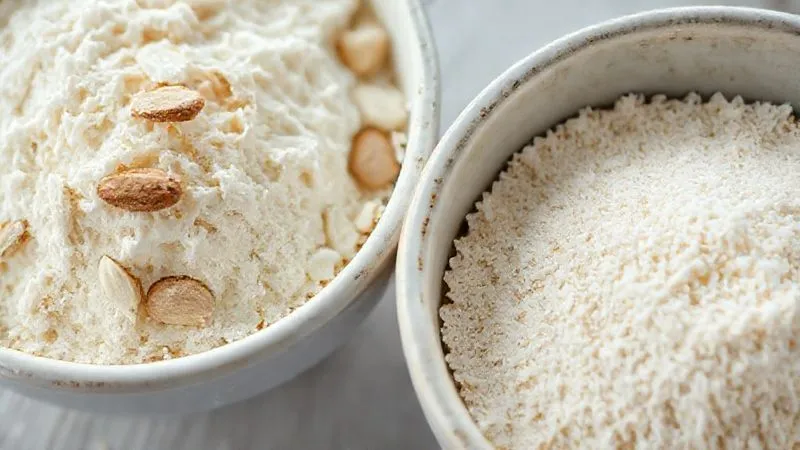 Photorealistic close-up of two bowls: one with light, fluffy almond flour, and another with fine, white wheat flour. The almond flour bowl should have a slightly coarser, more textured appearance, while the wheat flour is very fine. Cinematic lighting, sharp focus, depth of field, 8K hyper-detailed, professional photography.